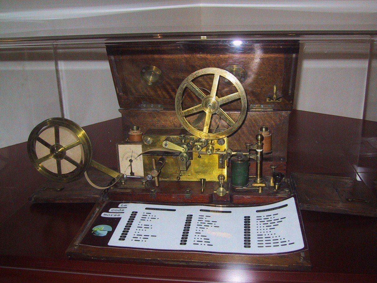 A photograph of one of Samuel Morse's early telegraph machines: a small wooden frame containing brass coils, a clockwork mechanism, and a strip of paper tape for recording dots and dashes.