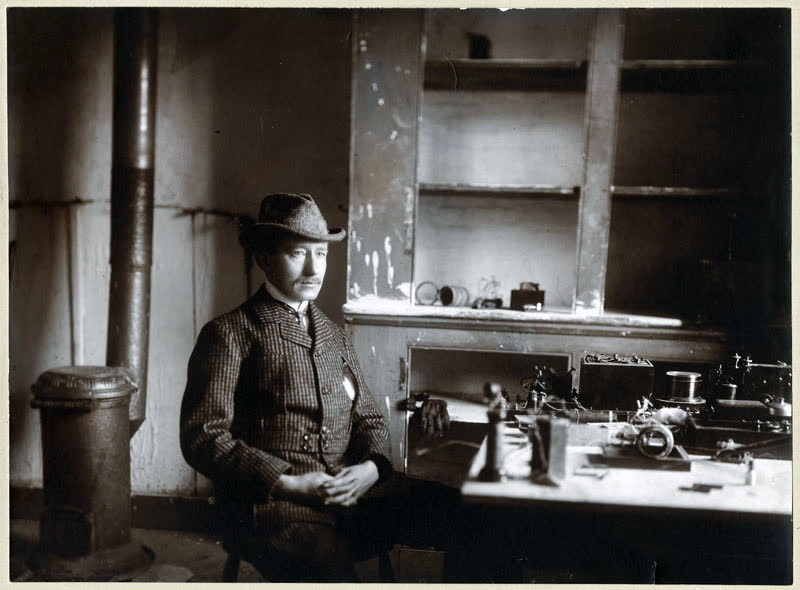 A black-and-white period photograph of Guglielmo Marconi seated inside Cabot Tower in Newfoundland, hunched over his wireless receiving apparatus — a wooden table covered in coils, knobs, batteries, and a strip of paper tape.