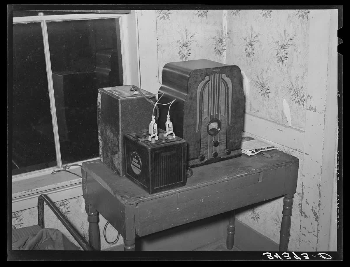 A 1939 documentary photograph of a small battery-powered wooden radio set sitting on a shelf inside a rural American farmhouse. Family possessions are visible around it.