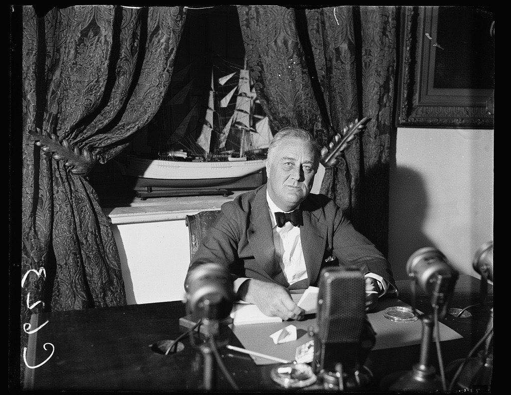 A black-and-white photograph of Franklin D. Roosevelt seated at a microphone, leaning forward to read from a typed script. Several broadcast network microphones cluster on the desk in front of him.