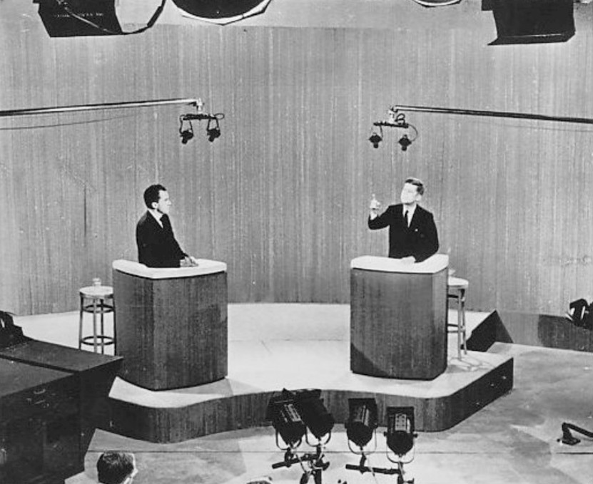 A black-and-white press photograph of John F. Kennedy and Richard Nixon standing at separate lecterns during a televised presidential debate. Both wear dark suits; a moderator's table sits between them.