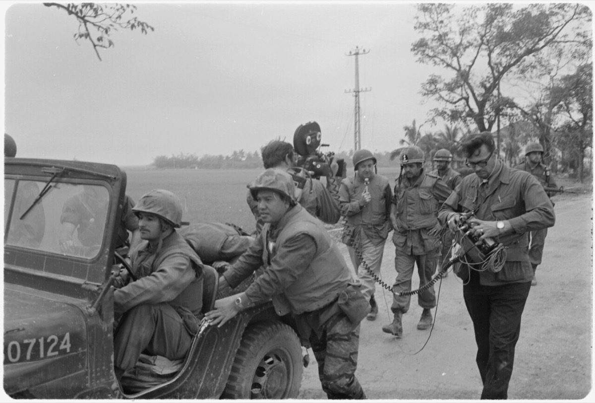 A black-and-white photograph showing CBS News anchor Walter Cronkite, wearing combat fatigues and a steel helmet, conducting an interview with a US Marine officer. A camera operator films from beside a jeep being used as a mobile dolly.