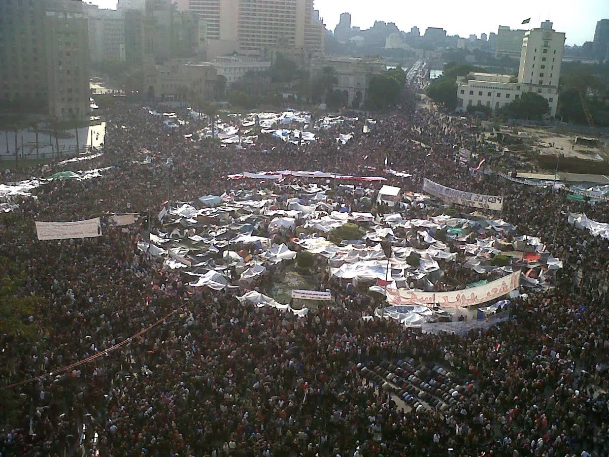 An aerial photograph of Tahrir Square in central Cairo, filled with hundreds of thousands of protesters. Egyptian flags are visible throughout the crowd; tall apartment buildings frame the square.