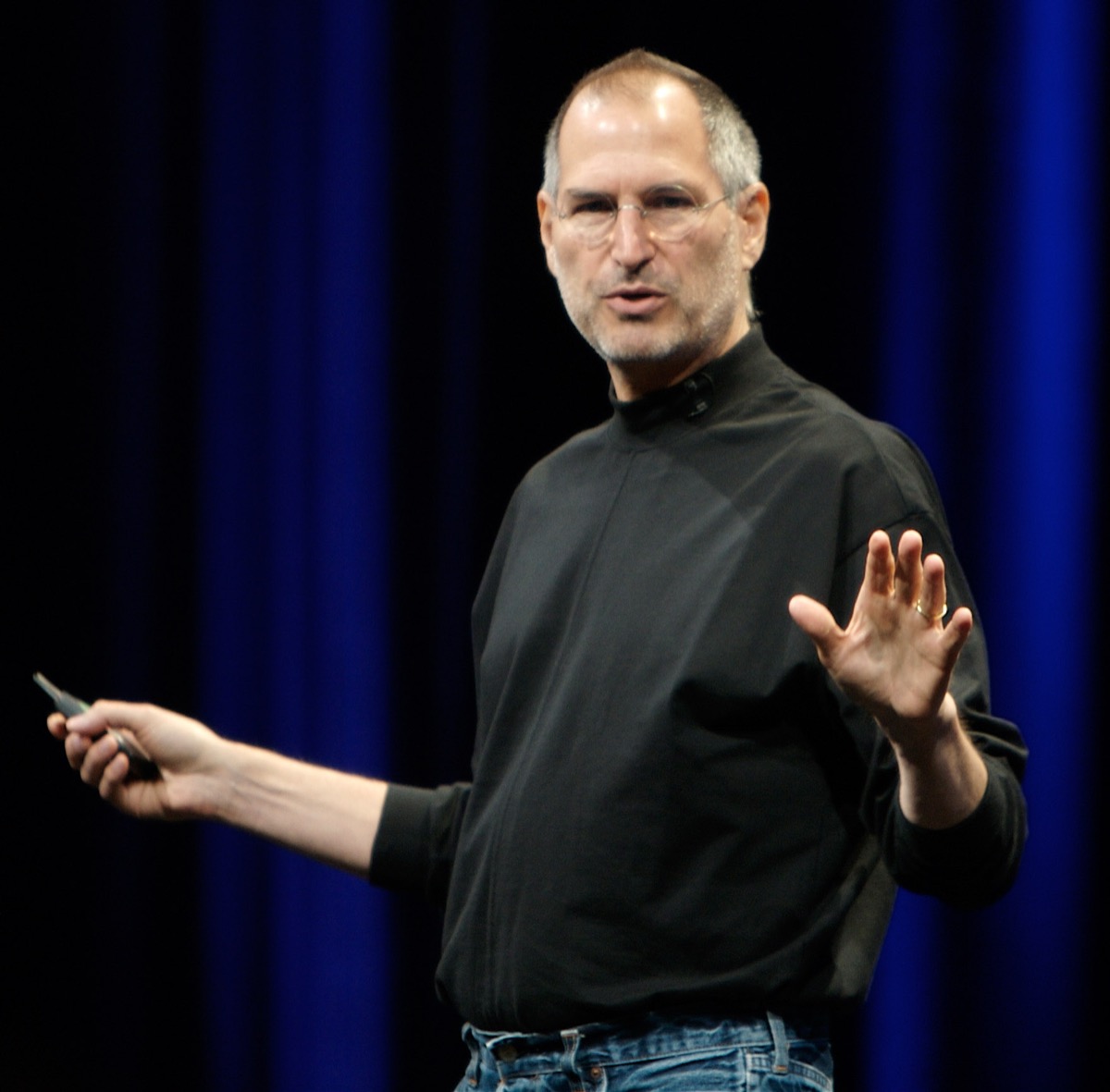 A 2007 colour photograph of Steve Jobs standing on stage in his trademark black turtleneck and jeans, gesturing toward a giant screen behind him. He is mid-presentation at an Apple conference.